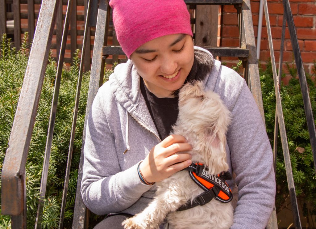 Maddie is sitting, looking down at her small dog, Cody. Maddie wearing a pink hat and grey sweater. Cody is wearing an orange vest that says "service dog". 