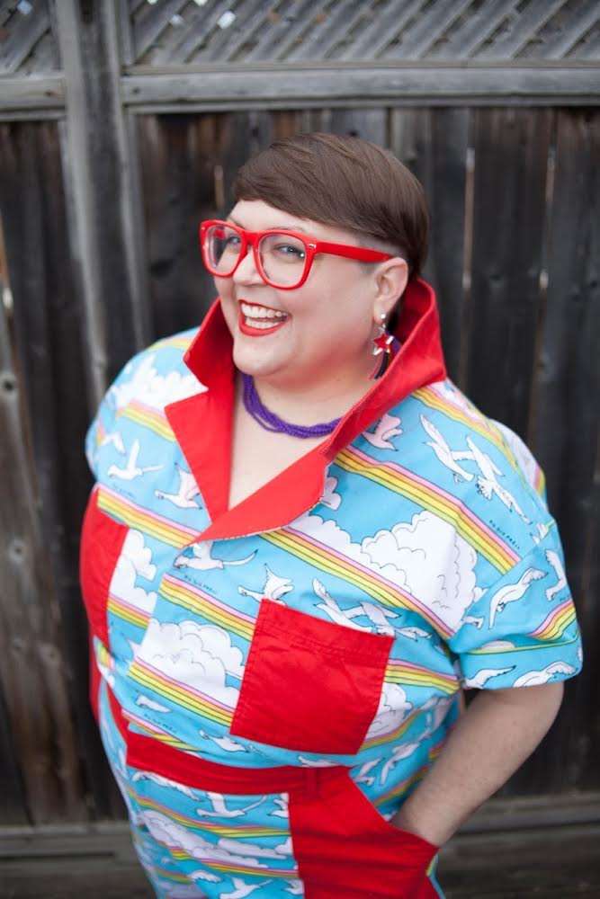 Sookie is smiling at the camera. They are wearing red glasses and a colourful jumpsuit. She is standing in front of a wood fence.