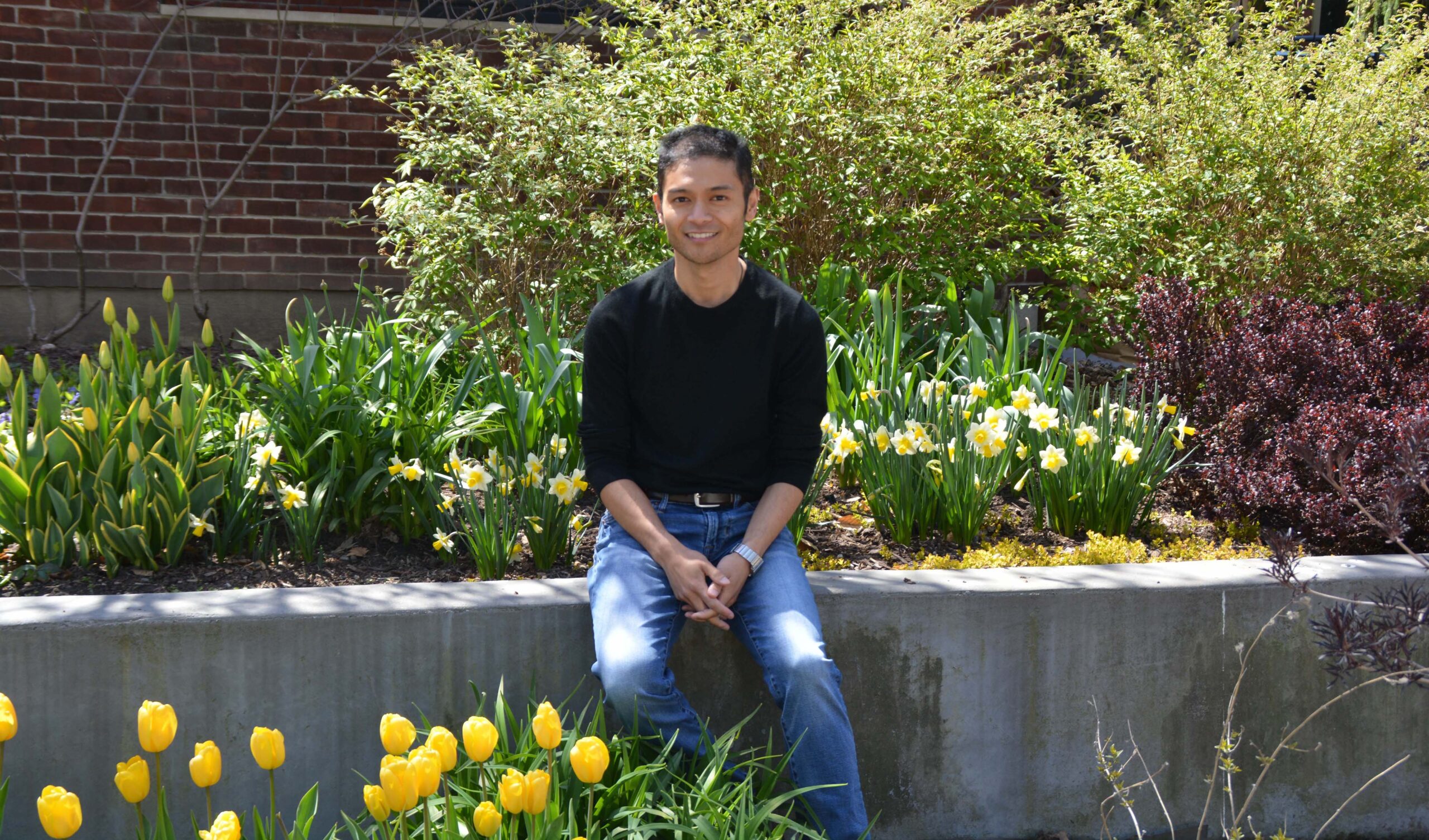 Jeffrey is sitting on a ledge and looking at the camera, smiling. He is wearing jeans and a black shirt. There are flowers/plants behind him and by his feet