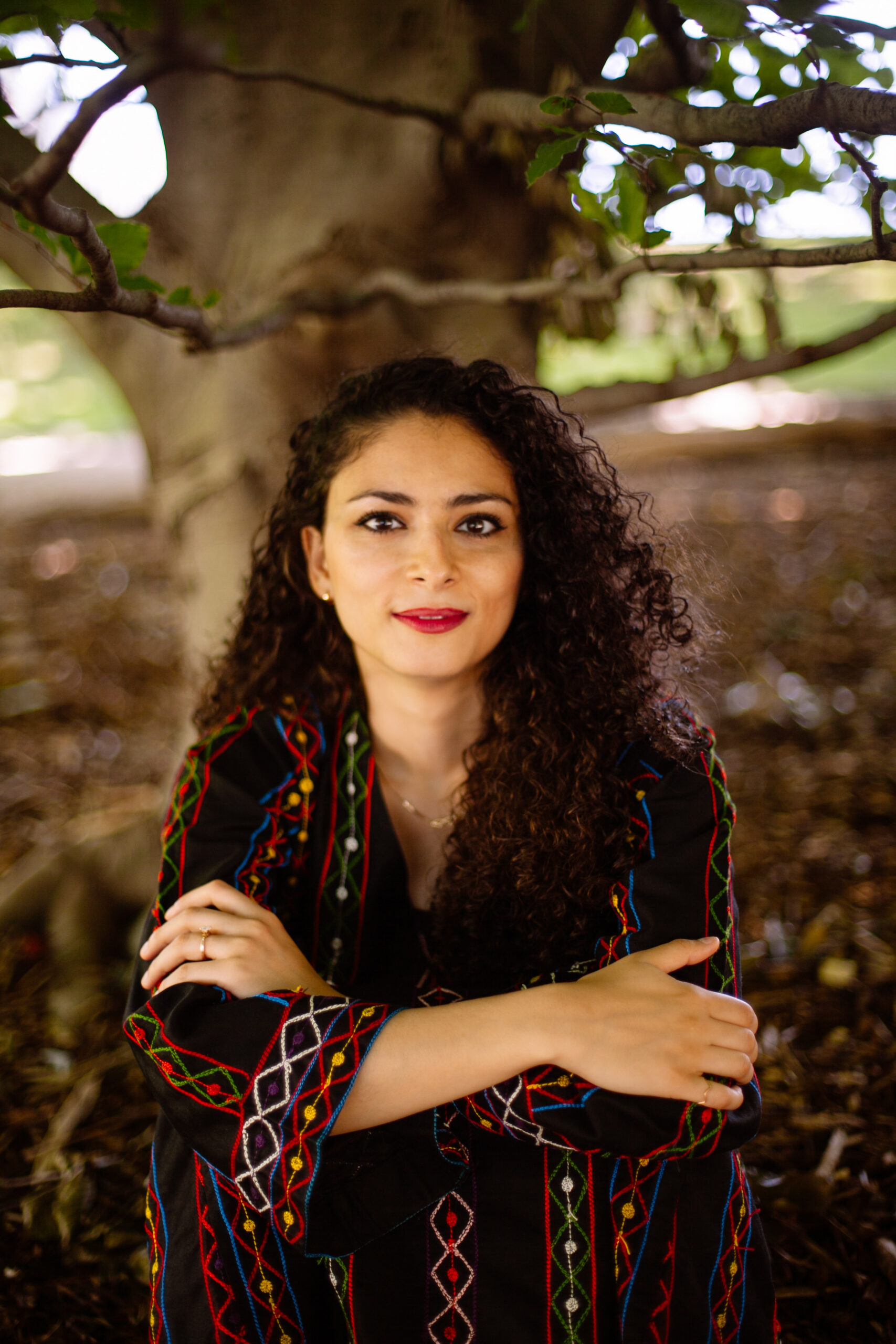 Alexandra is looking at the camera, smiling. Her arms are crossed and she is wearing a black jacket with a colourful pattern. There is a tree behind her.