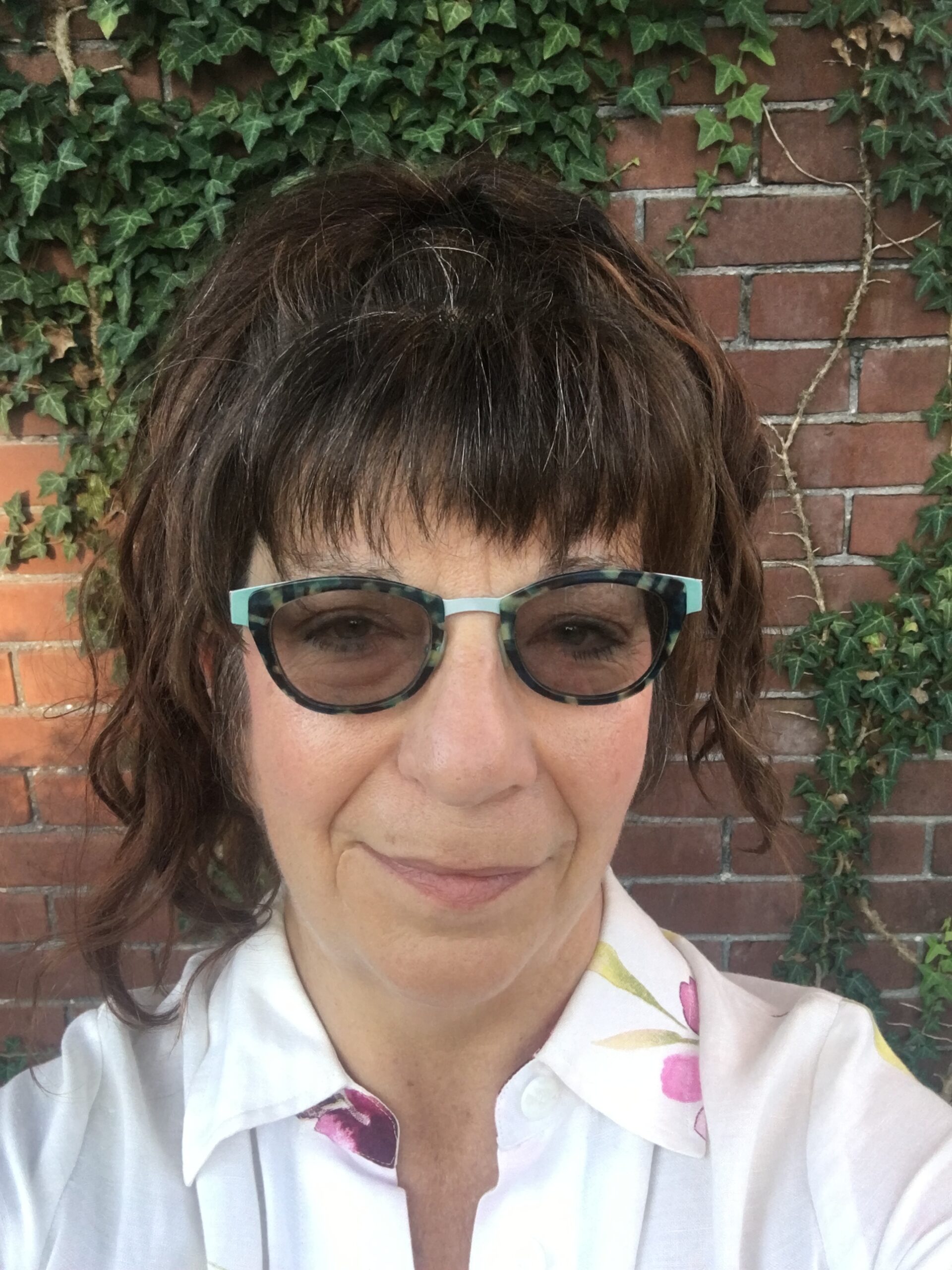Margaret is looking at the camera and smiling. She is wearing glasses and a white shirt. There is a brick wall with vines behind her.