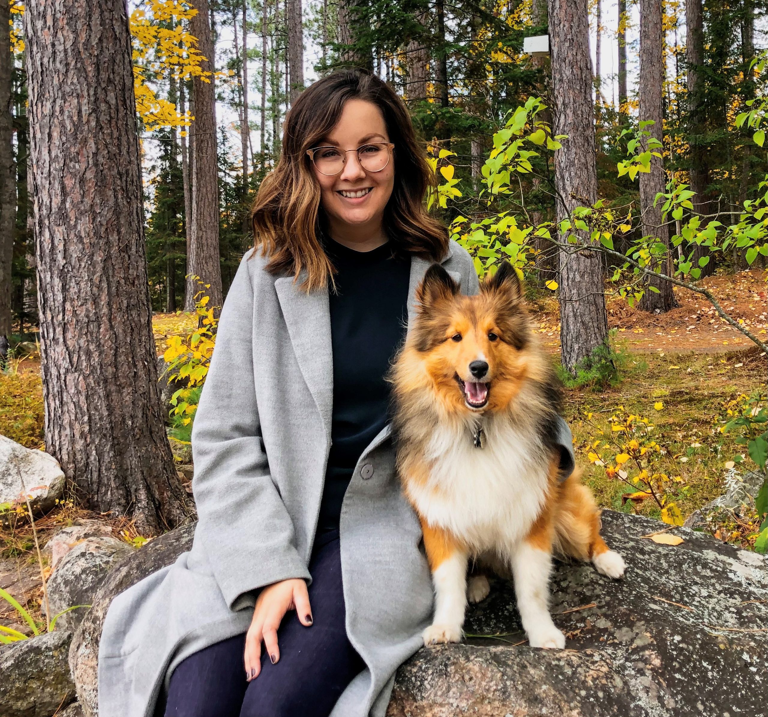 Sara is wearing a grey coat and is sitting on a rock, in front of trees, with her dog. Sara is smiling into the camera.