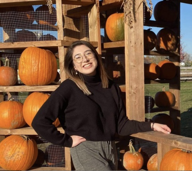 Colleen is wearing a black shirt and smiling into the camera. Colleen is standing in front of pumpkins.